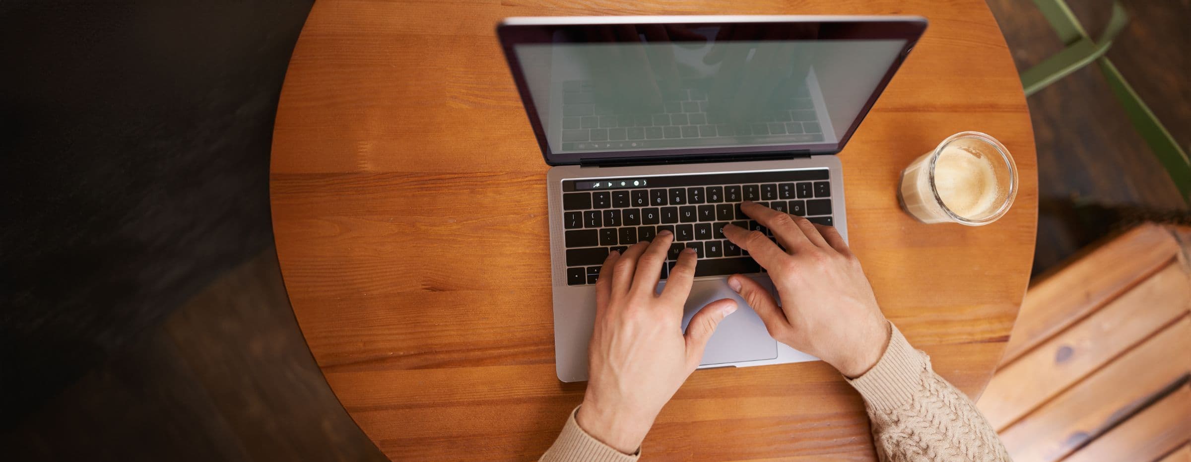 Top-down view of hands typing on a laptop at a wooden café table beside a latte